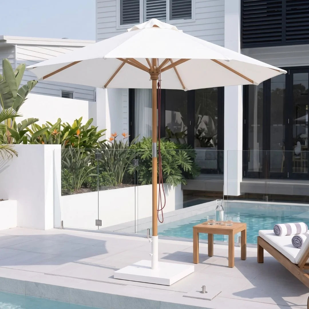 A large white patio umbrella with a natural wood pole and base stands beside a swimming pool on a modern outdoor deck. A small wooden side table with a bottle and glasses is nearby, along with a lounge chair and tropical plants in the background.