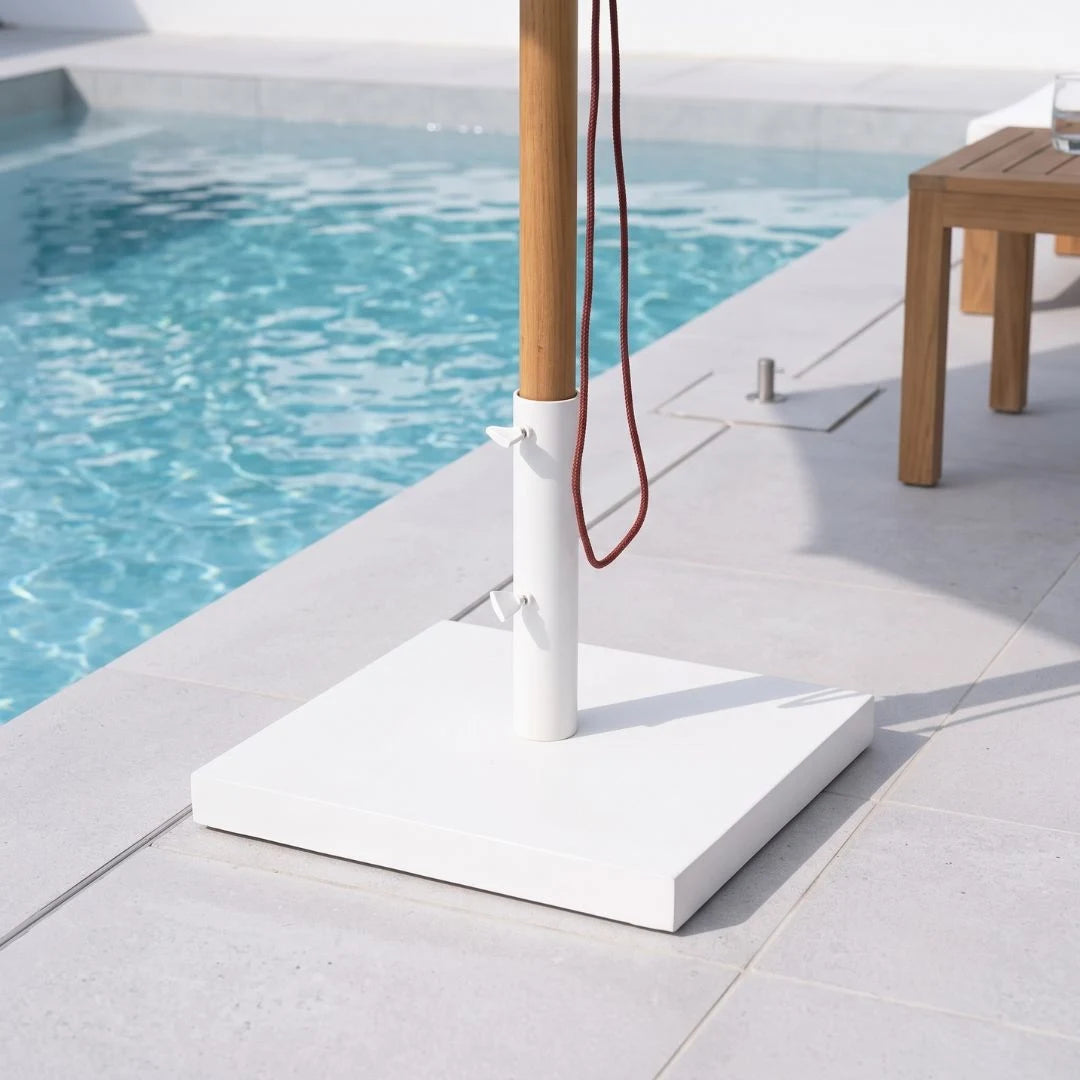 A white square patio umbrella base with a natural bamboo pole and red hanging cord, set on a light stone deck next to a swimming pool.