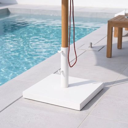 A white square patio umbrella base with a natural bamboo pole and red hanging cord, set on a light stone deck next to a swimming pool.