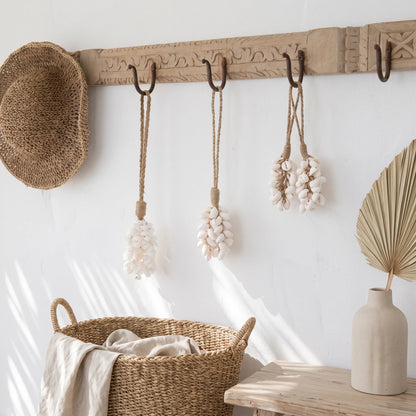 A carved wooden wall hook rack with dark metal hooks hangs in a bright, sunlit room. A woven straw hat hangs on the left, and three woven rope ornaments with shell-like white decorations hang from the center and right hooks.
