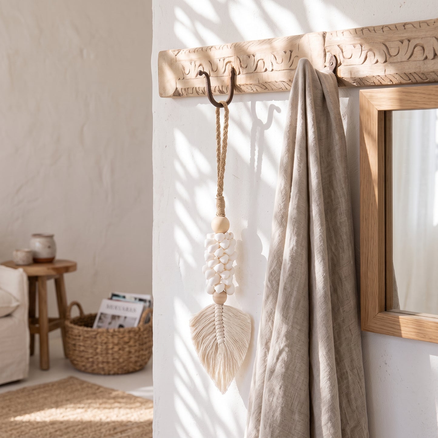 A beige woven wall hanging with a wooden bead center and a large teardrop-shaped tassel is suspended from a carved wooden entryway hook. A draped linen throw and part of a mirror are visible in a softly lit, neutral-toned interior.
