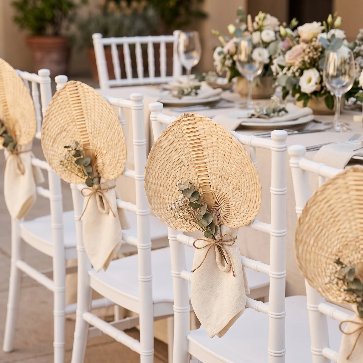 White wooden dining chairs decorated with woven straw fans and cream napkins, arranged around a floral wedding table setting.