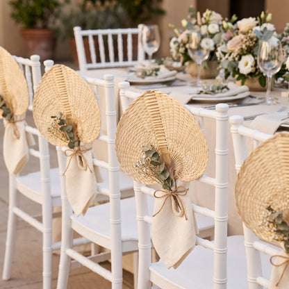 White wooden dining chairs decorated with woven straw fans and cream napkins, arranged around a floral wedding table setting.