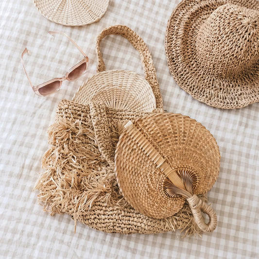 A styled flat-lay of natural woven straw accessories on a beige-and-white gingham fabric background, including a large fringed shoulder bag, a round woven hat, a smaller woven hat, a folding straw fan, and pink-tinted sunglasses.
