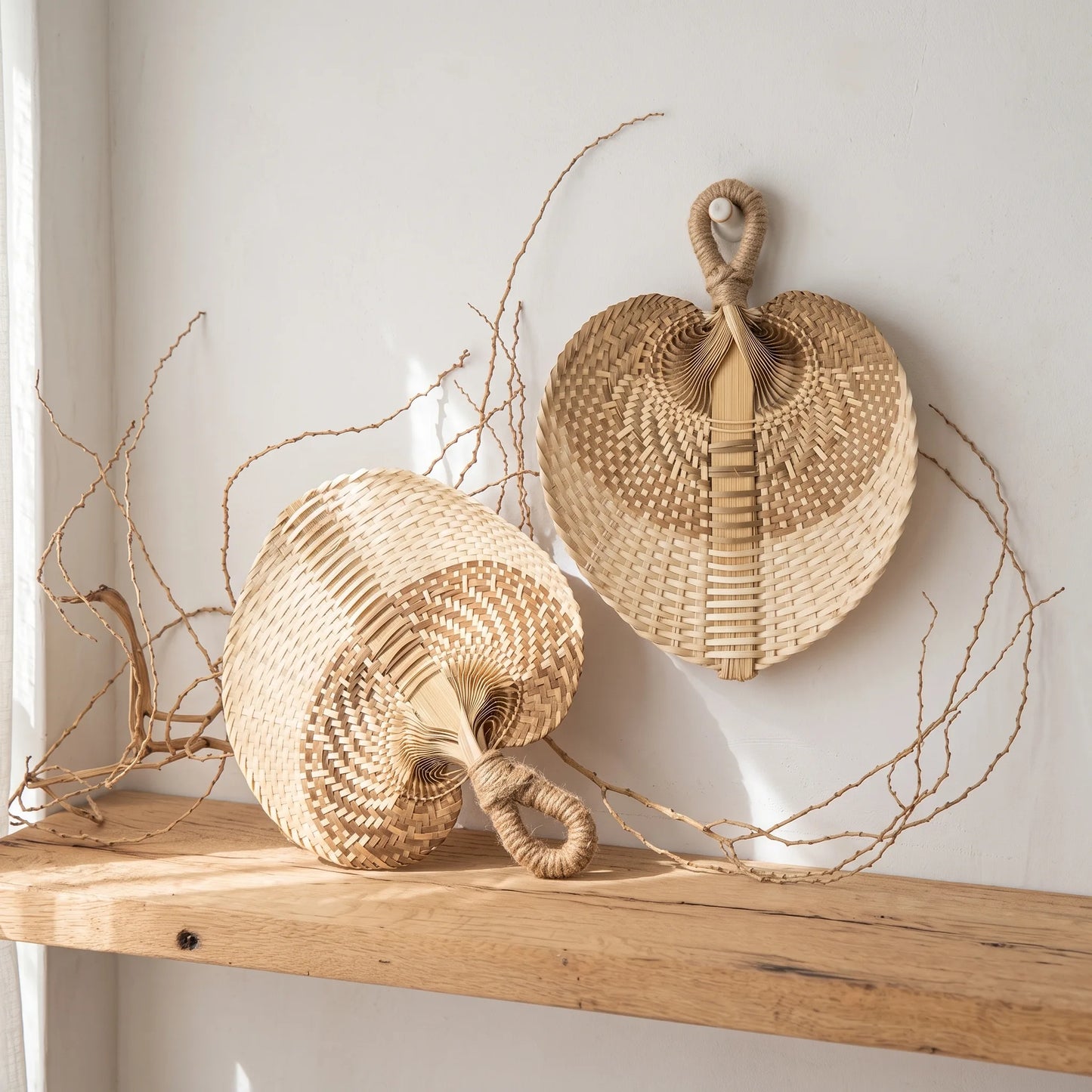 Two woven natural-fiber decorative baskets with looped handles, one standing upright and one lying on its side, displayed on a wooden shelf against a white wall with dried twigs around them.