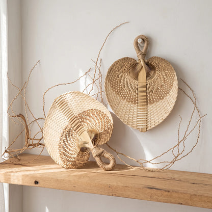 Two woven natural-fiber decorative baskets with looped handles, one standing upright and one lying on its side, displayed on a wooden shelf against a white wall with dried twigs around them.