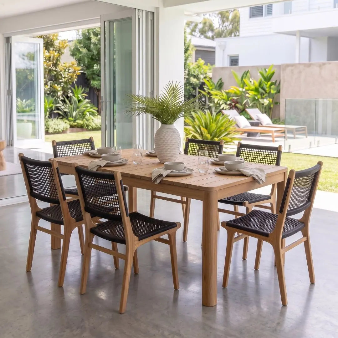 A rectangular wooden dining table with six woven-back chairs, set in a bright, modern outdoor patio dining area with plants and glass doors in the background.
