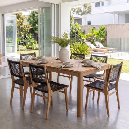 A rectangular wooden dining table with six woven-back chairs, set in a bright, modern outdoor patio dining area with plants and glass doors in the background.