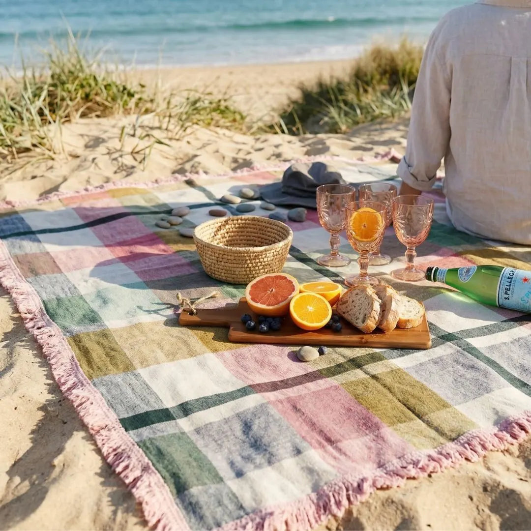 Person having a picnic on a colorful blanket by the beach with fruits and drinks.