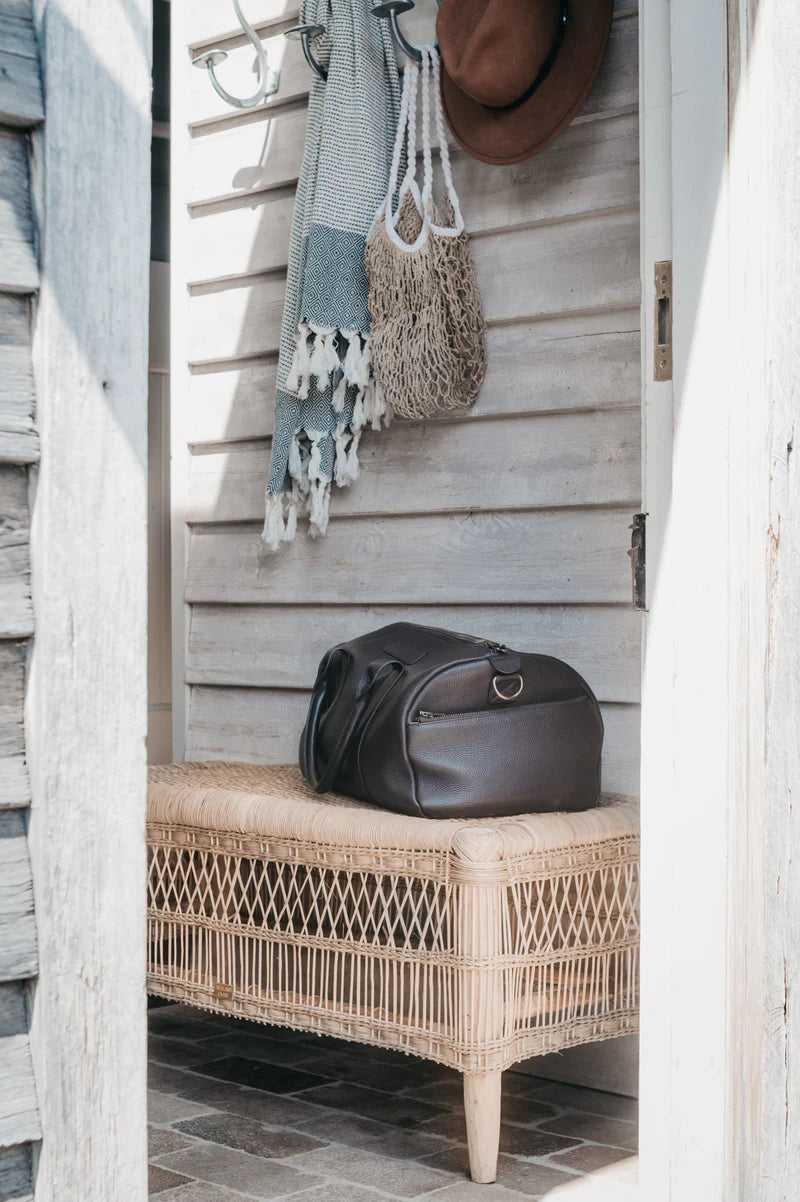 Brown leather bag on a wicker bench with a wooden wall and hanging items in the background