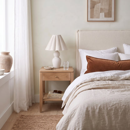 A styled bedroom scene featuring a light wood nightstand with a white ceramic table lamp, set beside a bed with neutral linens and a rust-colored accent pillow in a softly lit room.