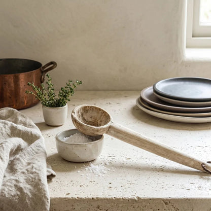 Wooden ladle on a stone surface with a pot, plates, and plant in the background.