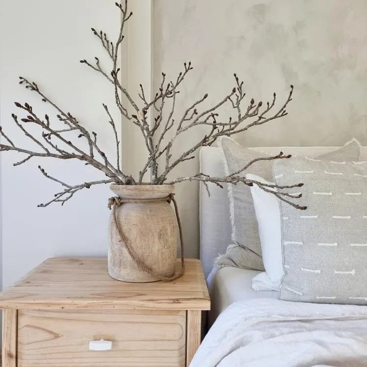 Decorative branches in a woven vase on a wooden nightstand next to a bed with patterned pillows.