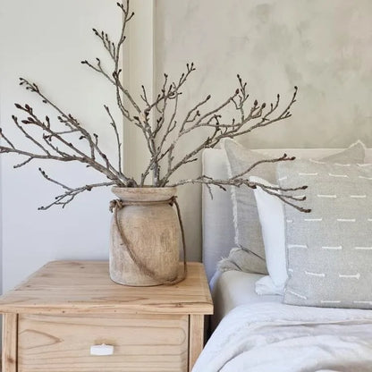Decorative branches in a woven vase on a wooden nightstand next to a bed with patterned pillows.
