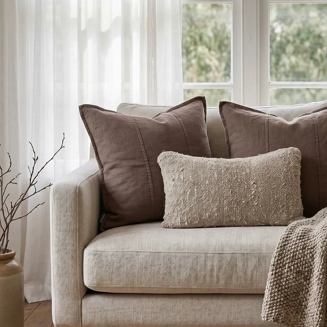 Beige sofa with brown and textured pillows in a room with white curtains and greenery outside.