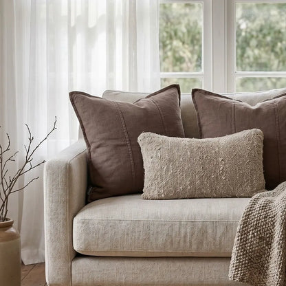 Beige sofa with brown and textured pillows in a room with white curtains and greenery outside.