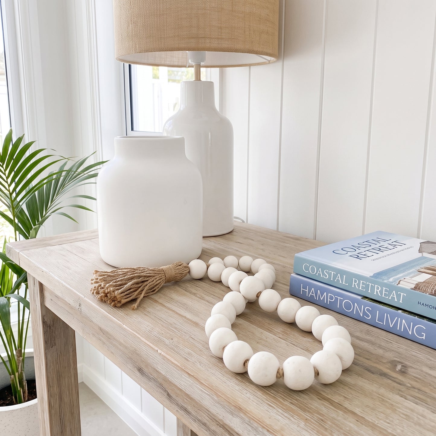 A white wooden bead garland with a tassel is styled on a light wood console table next to a white ceramic vase, a table lamp, and a stack of books in a bright, modern room.