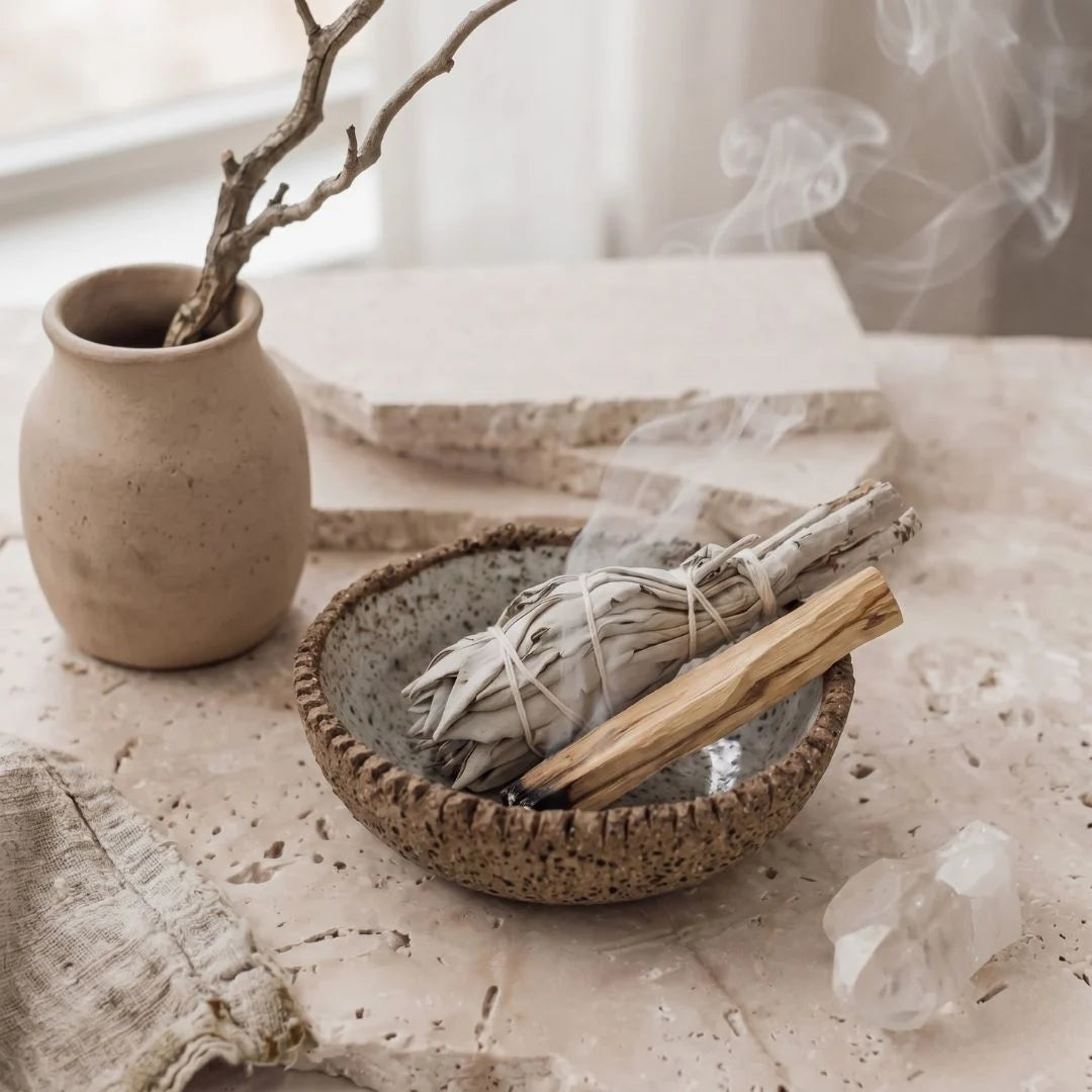 Ceramic bowl with smoldering sage sticks, crystal, and vase on a textured surface.