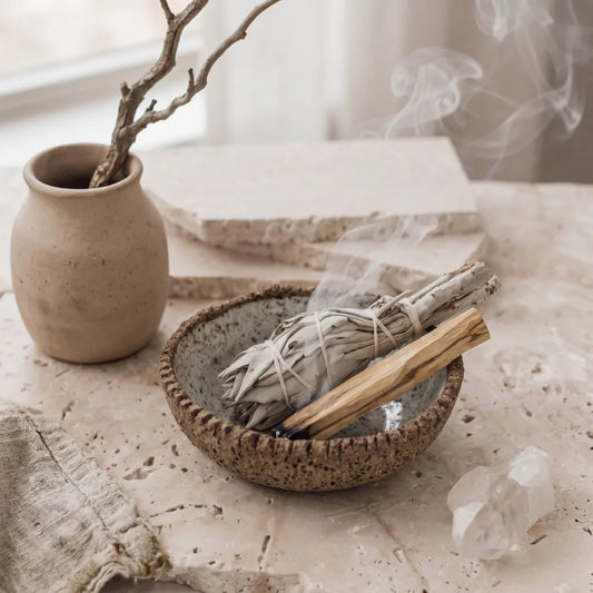 Ceramic bowl with smoldering sage sticks, crystal, and vase on a textured surface.