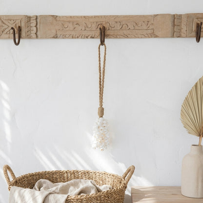 A boho-style woven rope tassel ornament with a white shell bead cluster hangs from a carved wooden wall rack with dark metal hooks. The scene is styled with a wicker basket and a ceramic vase on a light wood table against a white wall with soft window shadows.