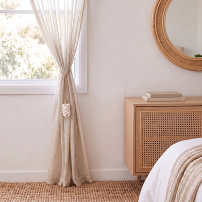 A beige linen curtain tied back from a window in a bright, minimalist bedroom with a wooden cabinet, woven rug, and round mirror.