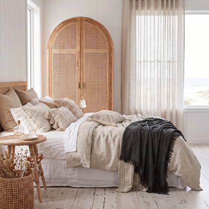 A styled bedroom scene featuring a bed dressed in neutral beige and white linens with a dark charcoal throw blanket, set against a light wood arched headboard with woven cane panels. The room has white paneled walls, sheer curtains, and a rustic side table with dried flowers.