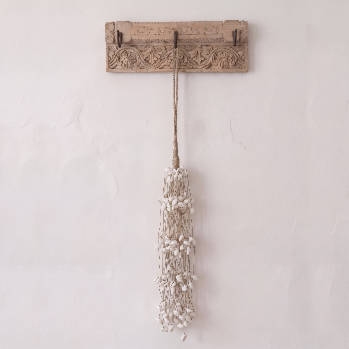 A rustic carved wooden wall hook with two hooks, hanging a long tassel made of beige cord and small white beads, against a textured off-white wall.