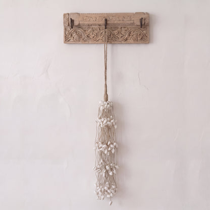 A rustic carved wooden wall hook with two hooks, hanging a long tassel made of beige cord and small white beads, against a textured off-white wall.