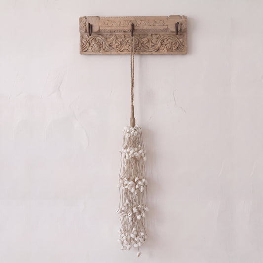 A rustic carved wooden wall hook with two hooks, hanging a long tassel made of beige cord and small white beads, against a textured off-white wall.