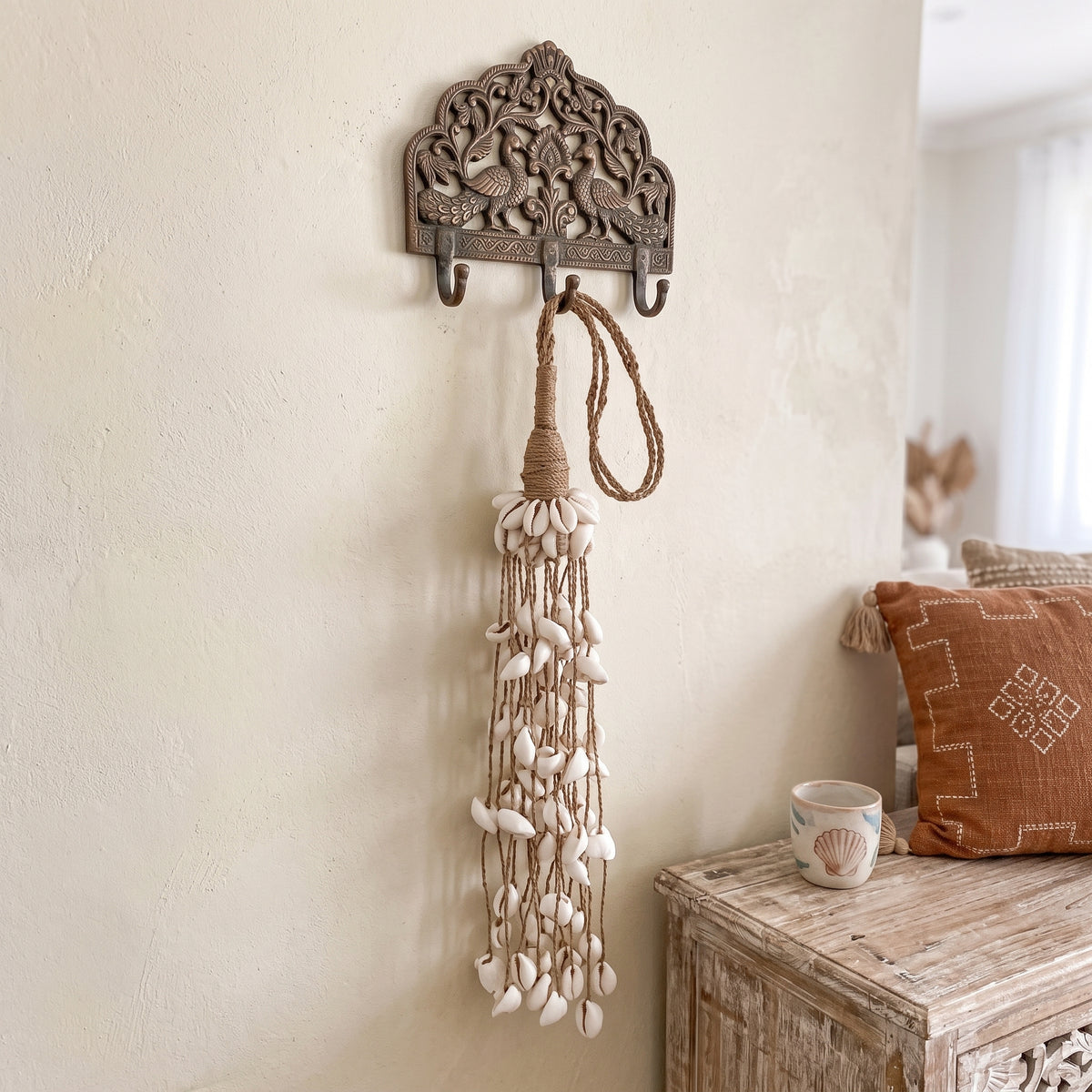 A bohemian-style wall hook holder featuring an ornate carved metal top with peacock motifs and a long tassel made of natural rope and white seashells, hanging on a textured cream wall next to a rustic wooden side table.