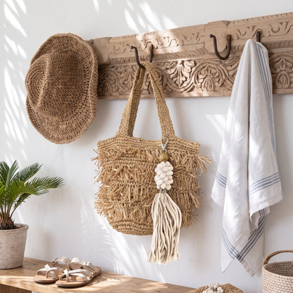 A natural woven straw tote bag with fringe and a tassel hangs from a carved wooden wall rack. A matching straw hat is hanging on the left, a white towel with blue stripes hangs on the right, and a small potted plant and sandals sit on a wooden bench below.