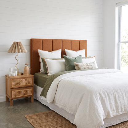 A styled bedroom scene featuring a bed with a tall, padded rust-orange upholstered headboard, dressed in white linens and green accent pillows, next to a light wood nightstand with a pleated beige lamp.