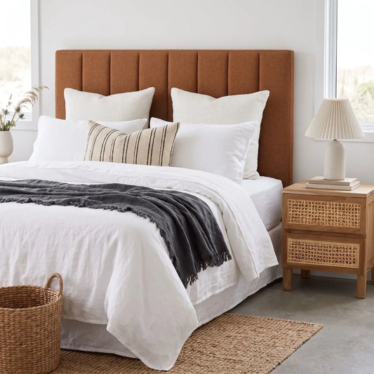 A neatly made bed with white bedding, a dark charcoal throw blanket, and a striped accent pillow, set against a prominent rust-brown upholstered headboard. A light wood nightstand with woven cane panels and a pleated table lamp sit on either side, with a woven basket on the floor in the foreground.