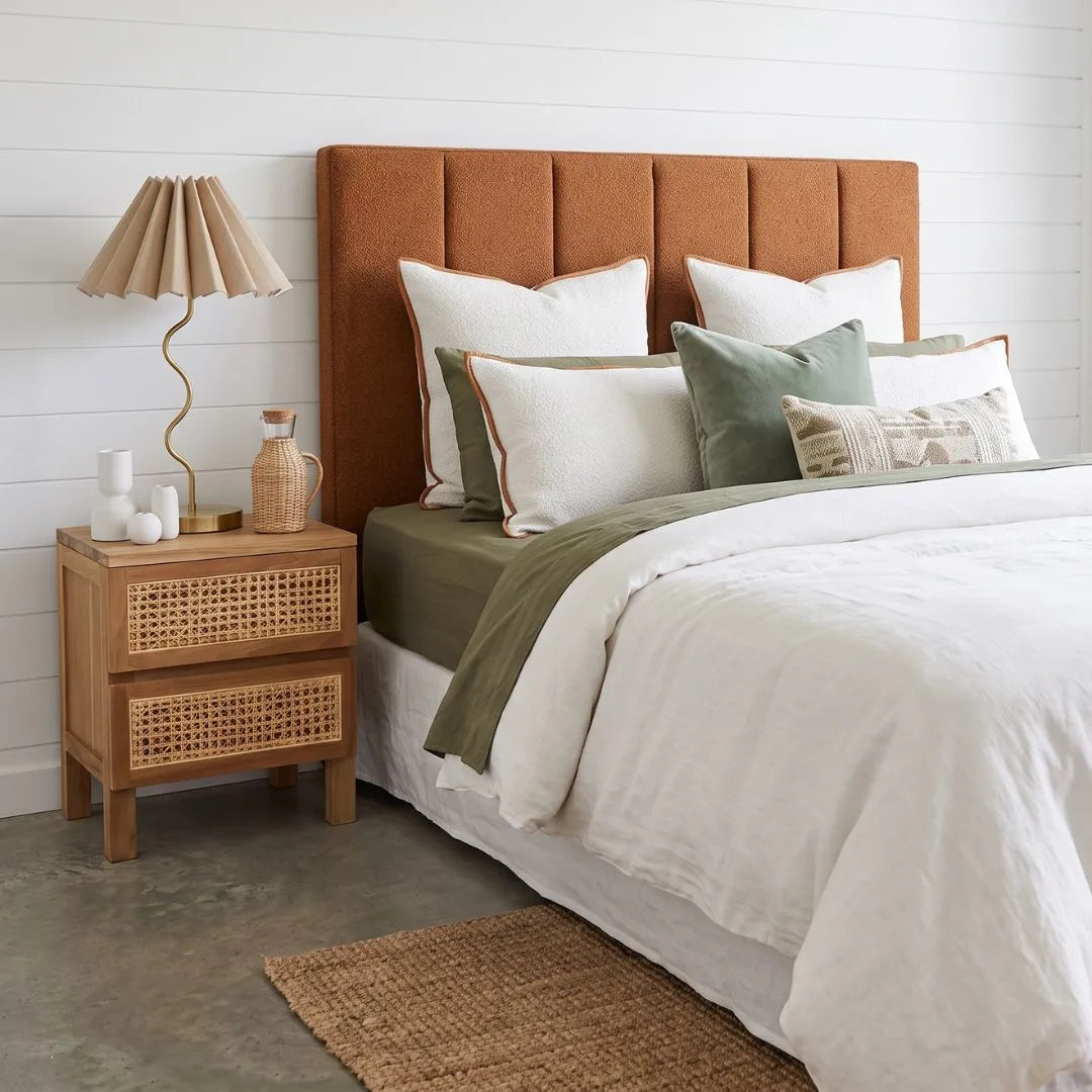 A styled bedroom scene featuring a bed with a tall, padded rust-orange upholstered headboard, dressed in white linens with sage green and olive accent pillows and a throw. A matching wooden cane-topped nightstand with a pleated beige table lamp sits on the left against a white shiplap wall.