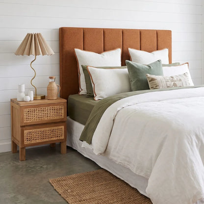 A styled bedroom scene featuring a bed with a tall, padded rust-orange upholstered headboard, dressed in white linens with sage green and olive accent pillows and a throw. A matching wooden cane-topped nightstand with a pleated beige table lamp sits on the left against a white shiplap wall.