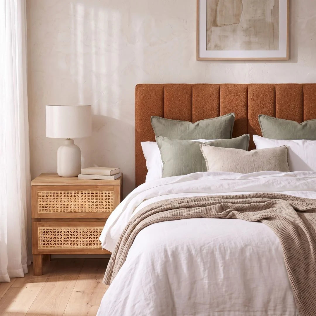 A styled bedroom scene featuring a bed with a tall, vertically channel-tufted rust-orange upholstered headboard, styled with white linens, sage green pillows, and a beige knit throw blanket. A matching rust-orange nightstand with woven cane panels stands on the left next to a white ceramic table lamp.