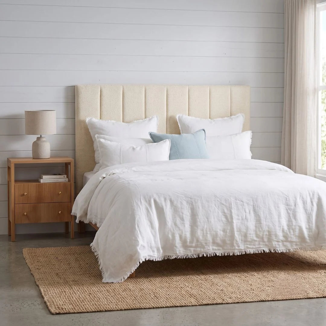 A neatly made bed with white bedding and pillows, set against a light wood-paneled wall with a beige upholstered headboard. A matching wood nightstand with a lamp sits on the left, and a woven jute rug covers the floor.