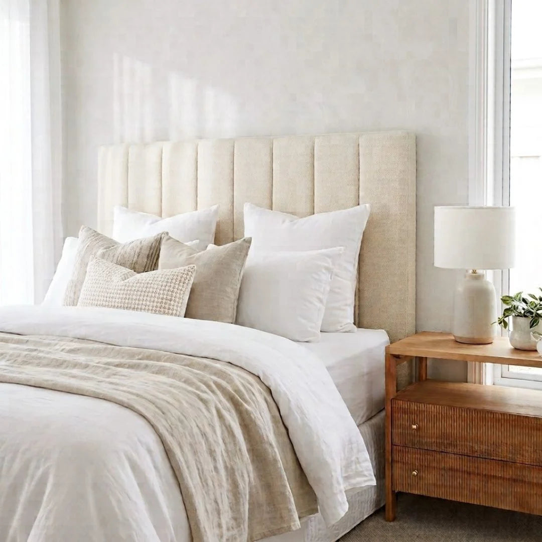 A styled bedroom scene featuring a bed with a tall, vertically channel-tufted upholstered headboard in a warm beige fabric. The bed is dressed in white linens, cream pillows, and a textured beige throw blanket.