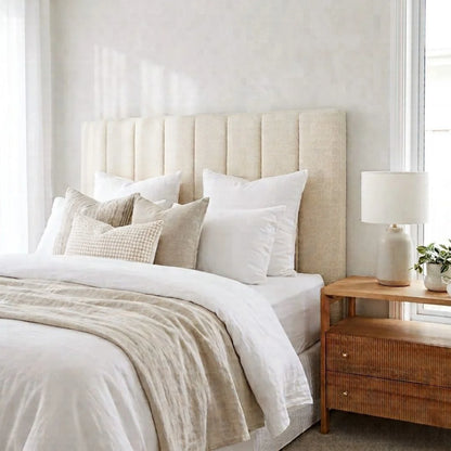 A styled bedroom scene featuring a bed with a tall, vertically channel-tufted upholstered headboard in a warm beige fabric. The bed is dressed in white linens, cream pillows, and a textured beige throw blanket.