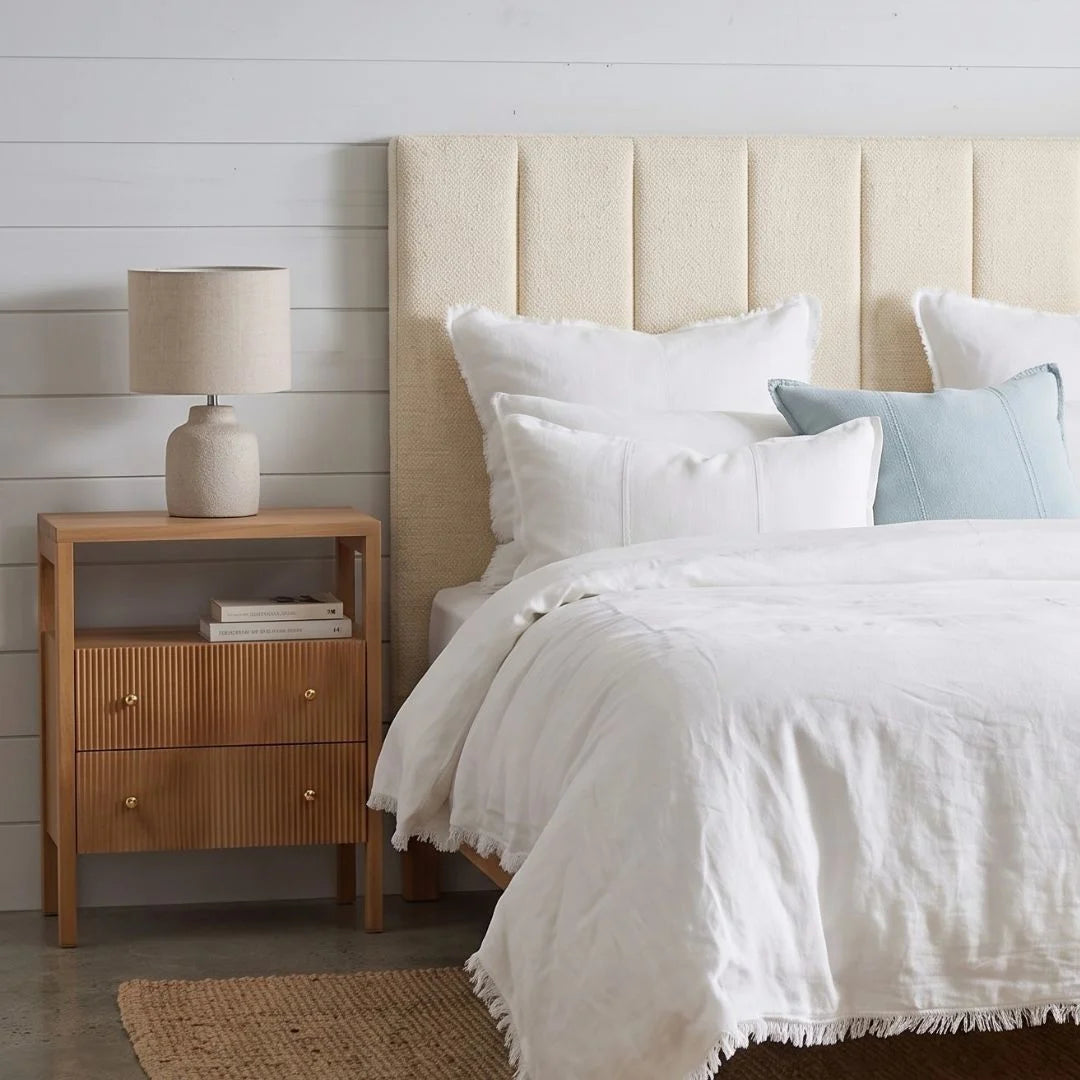A styled bedroom scene showing a bed with white bedding, a light beige upholstered headboard, a matching wood nightstand with a table lamp, and a pale blue accent pillow against a white shiplap wall.