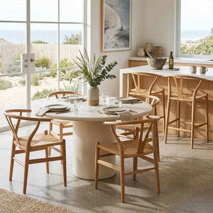 Dining area with a round table and wooden chairs in a bright room with large windows.