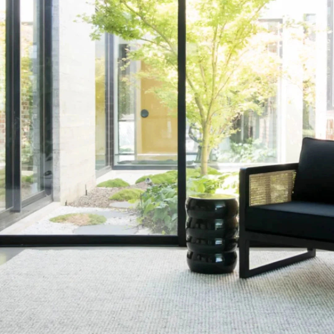 Modern interior with a black chair and a cylindrical black side table in front of large glass windows with a garden view.
