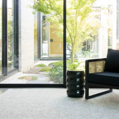 Modern interior with a black chair and a cylindrical black side table in front of large glass windows with a garden view.
