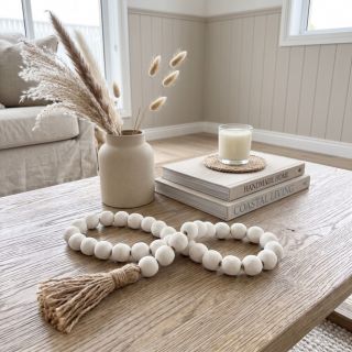 A decorative garland made of matte white round beads with small natural wood spacers and a jute tassel, draped over stacked books on a wooden stool in a softly styled living room.