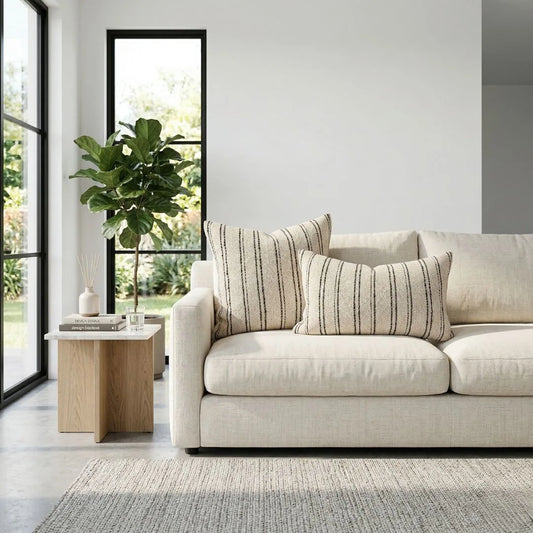 Beige sofa with striped cushions in a modern living room with large windows and a plant.