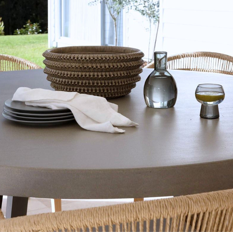 A round taupe-brown outdoor dining table set with a stack of woven brown bowls, dark gray plates, a white napkin, a clear glass decanter, and a small glass bowl, surrounded by woven wicker chairs.