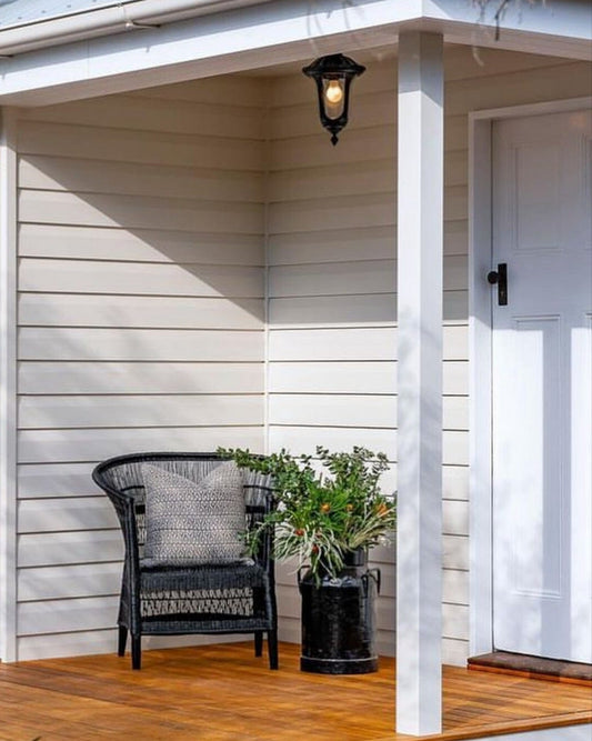 A black Malawi cane chair placed on a wooden deck outside a white house, with a potted plant beside it.