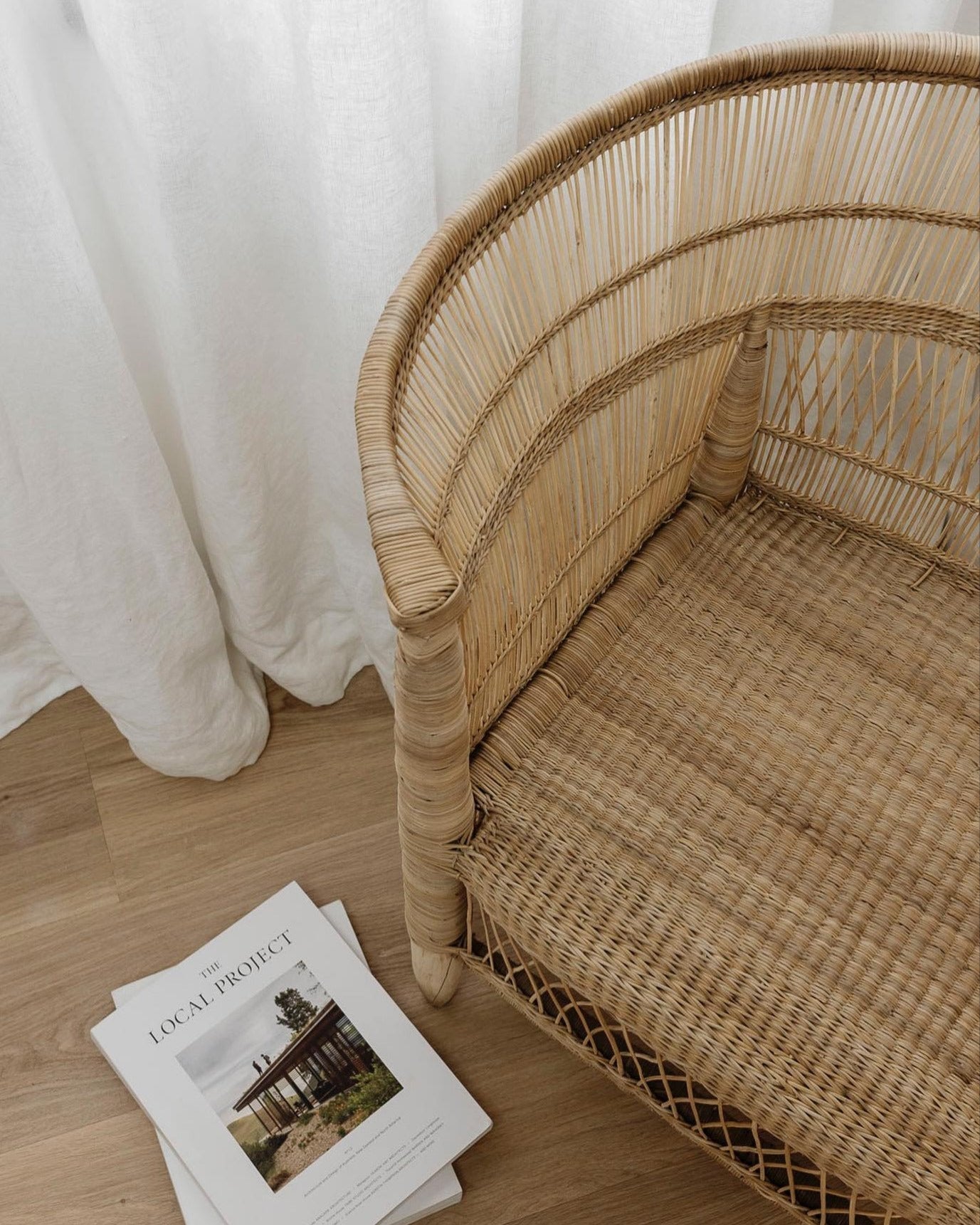Wicker chair with a stack of books on a wooden floor next to white curtains.
