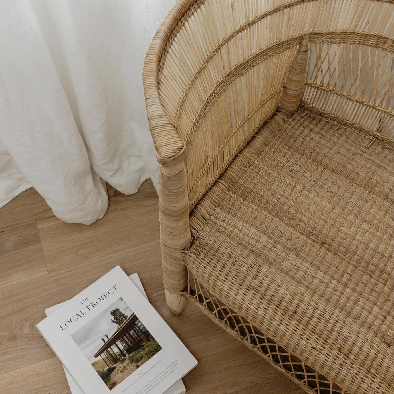 Close up of Malawi Cane Chair with a stack of books on a wooden floor next to white curtains.