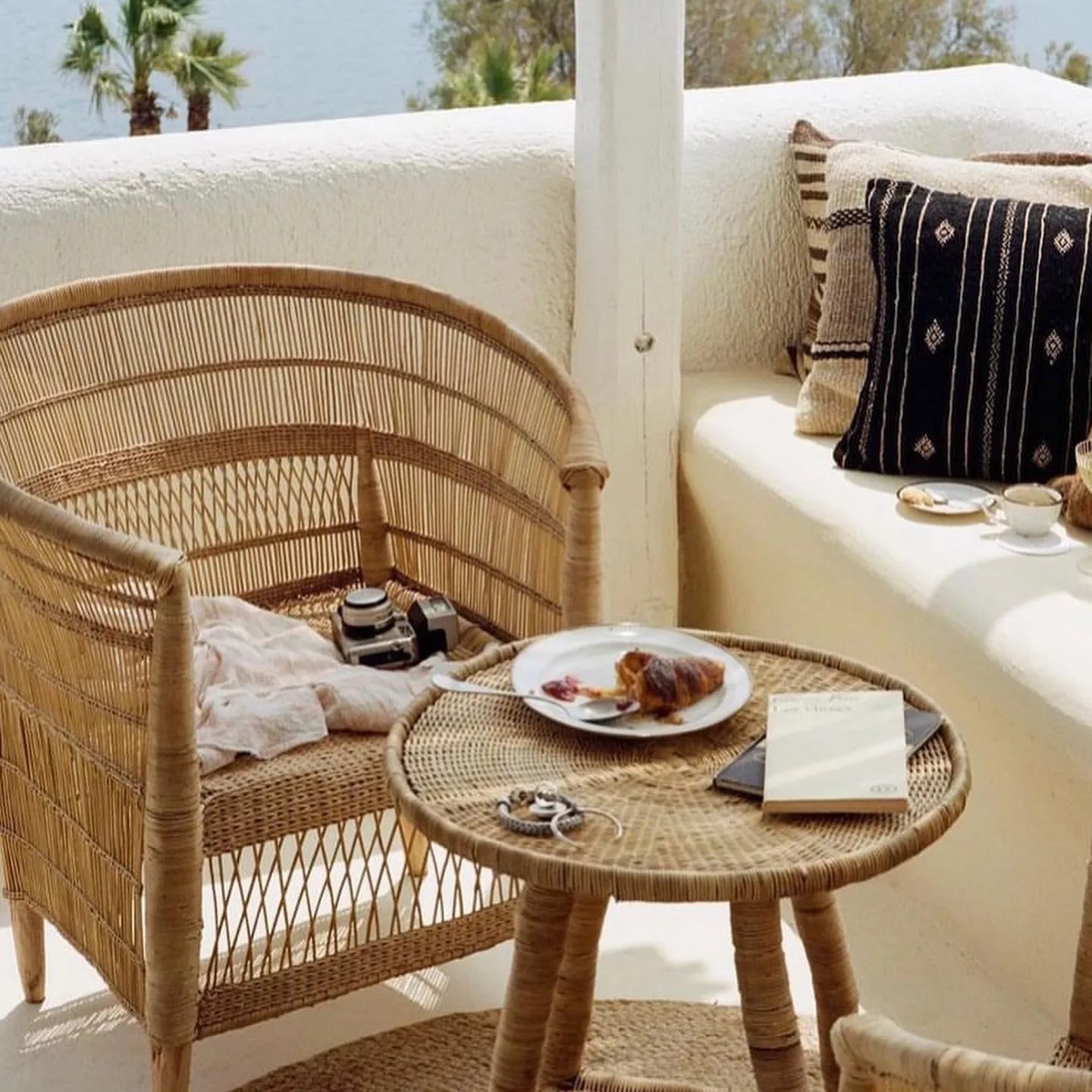 Wicker chair and table set on a balcony with cushions and a plate of food.